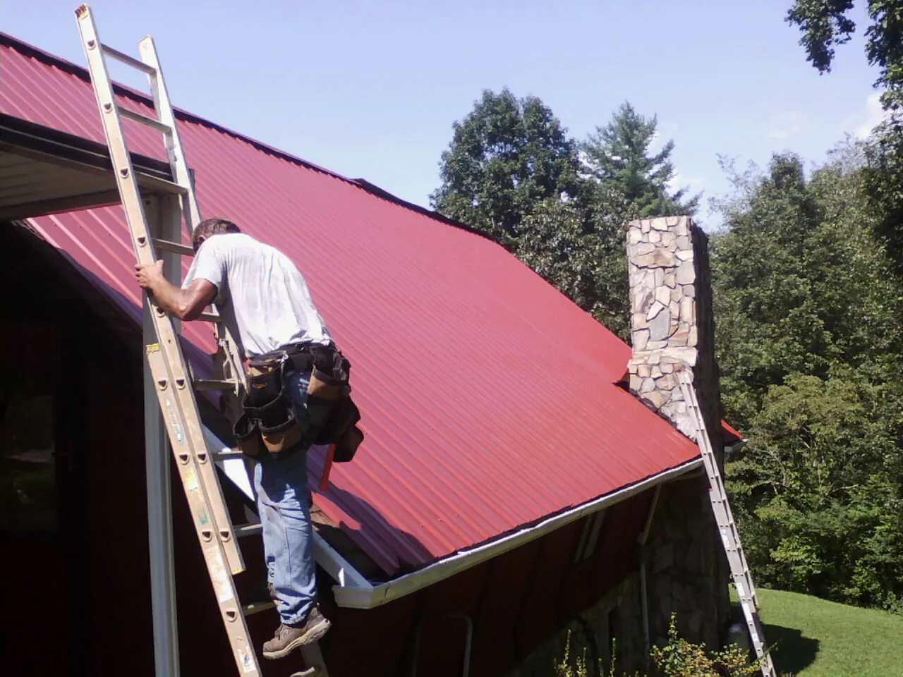 Roofer on a ladder beside a red metal roof and stone chimney — Soffit & Fascia Repair by Roofing Brothers in Truesdale